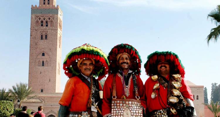 Trois vendeurs d'eau vêtus de façon traditionnelle posant devant la tour de la mosquée Koutoubia sous un ciel bleu.