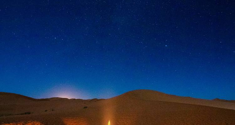 Ciel nocturne étoilé au-dessus de dunes de désert silencieuses avec un petit feu de camp qui brille à la base.