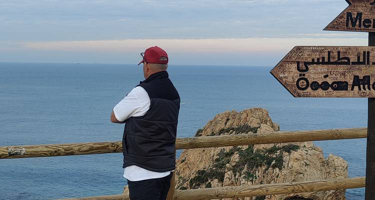 Homme en casquette contemple une mer bleue depuis un point de vue en bois avec des panneaux directionnels en arabe.
