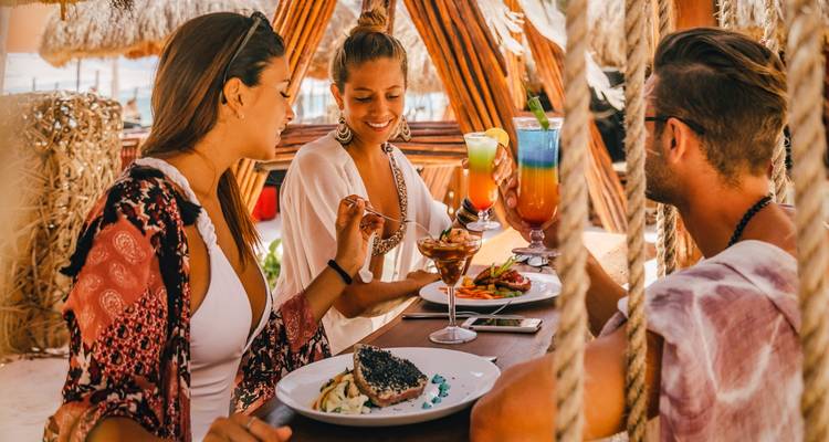 People enjoying a meal with colorful cocktails in a tropical setting.