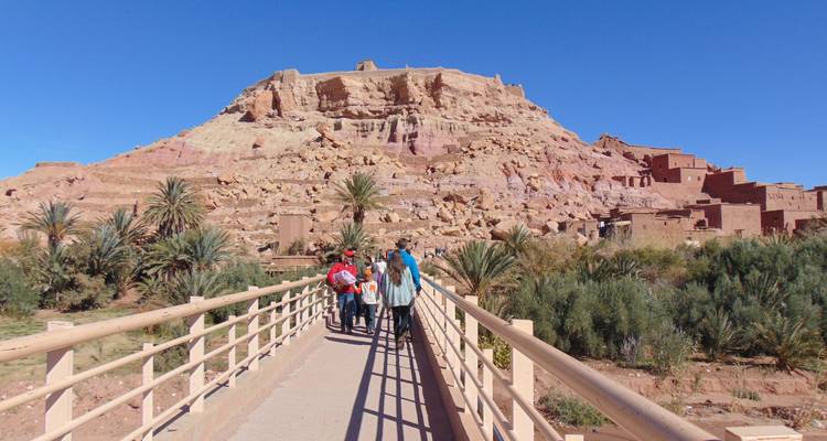Personas cruzando un puente peatonal hacia una gran colina con estructuras históricas.