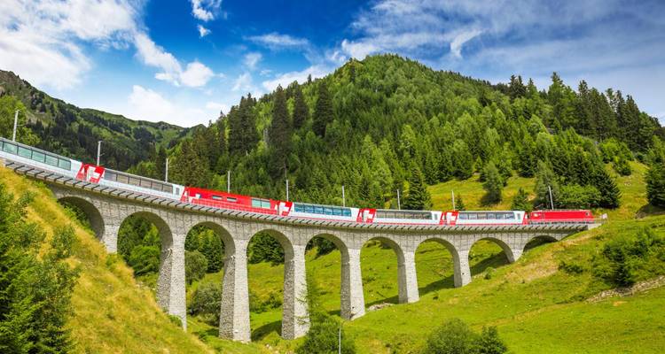Tren panorámico rojo y blanco cruzando un viaducto de piedra alto a través de un exuberante paisaje alpino verde.