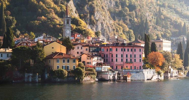 Pueblo colorido junto al lago con casas de colores pastel, una torre de reloj, y follaje otoñal reflejándose en las aguas tranquilas del lago.