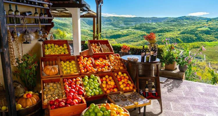 Puesto de productos al aire libre con cajas de manzanas de colores y botellas de vino local con vista a colinas ondulantes.