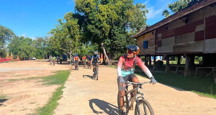 Une file de cyclistes traverse un village rural en passant devant des maisons en bois traditionnel et des arbres feuillus.