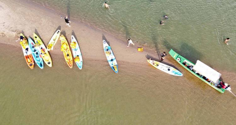 Vue aérienne de kayaks colorés alignés sur la rive sableuse d'une rivière avec des nageurs et un bateau à proximité