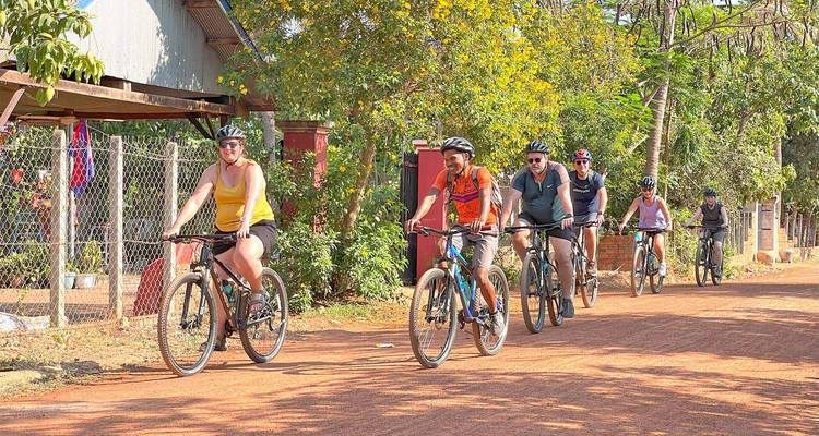 Un grupo de ciclistas pedalea por un camino rural polvoriento de color rojo, rodeado de simples casas de pueblo y árboles.