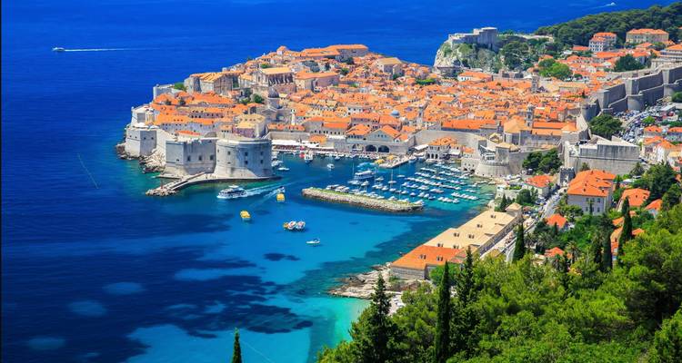 Panoramic aerial view of Dubrovnik’s walled old town with terracotta roofs surrounded by deep blue Adriatic waters and a marina.