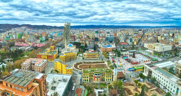 Wide cityscape of Tirana showing colorful government buildings and Skanderbeg Square under a dramatic cloudy sky.