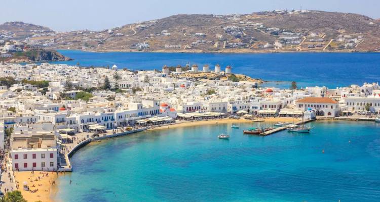 Curving bay of Mykonos town with white cube houses and famous hillside windmills beside clear Aegean waters.