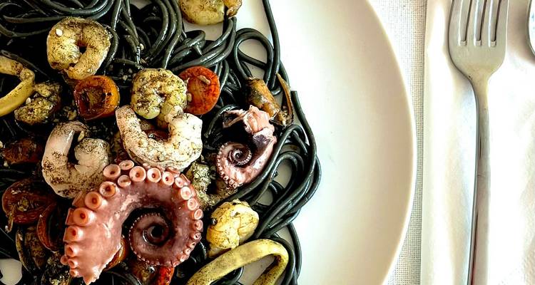 Close-up van zwarte inktvis-pastasaus gemengd met octopus, garnalen, inktvis en cherrytomaten op een wit bord.