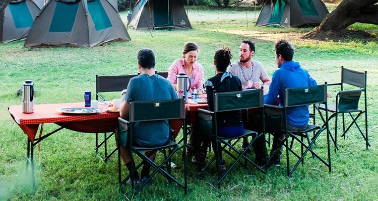 Los viajeros comparten una comida de campamento en mesas plegables al lado de tiendas de campaña armadas en las llanuras cubiertas de hierba.