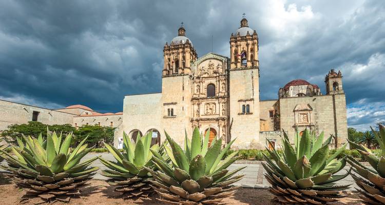 Die majestätische Kirche Santo Domingo de Guzmán in Oaxaca erhebt sich hinter stacheligen Agavenpflanzen unter stürmischem Himmel.
