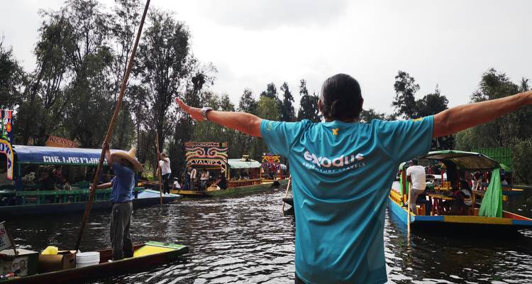 Reisender breitet Arme auf buntem Trajinera-Boot inmitten lebhafter Xochimilco-Kanäle aus, erfüllt von Musik und schwimmenden Gaststätten.