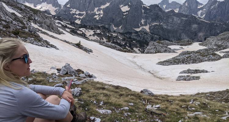Femme assise sur une pente de montagne enneigée avec des pics escarpés en arrière-plan.