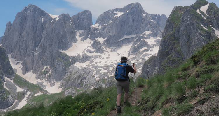 Hiker walking on a mountainous trail with snowy peaks.