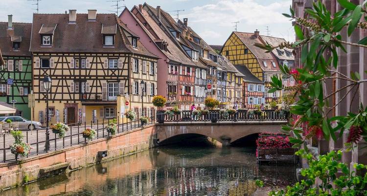 Half-timbered houses line a tranquil canal crossed by a flower-filled bridge.