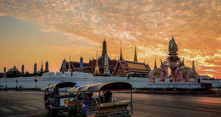 Tuk-tuks passeren het verlichte Grote Paleis en Erawan-standbeeld bij zonsondergang onder een dramatische oranje hemel in Bangkok.