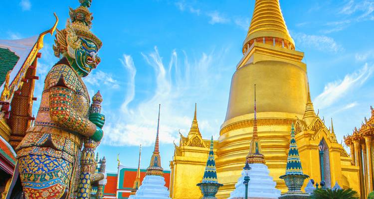 Levendige close-up van gouden stoepa's en een torenhoge Yaksha bewaker in Wat Phra Kaew onder een heldere blauwe hemel.