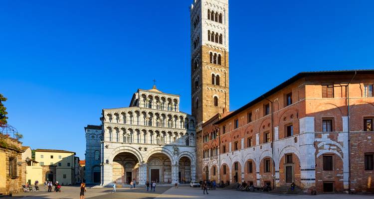 Sun-lit square fronting Lucca Cathedral with its ornate white façade and tall bell tower against a deep blue sky.