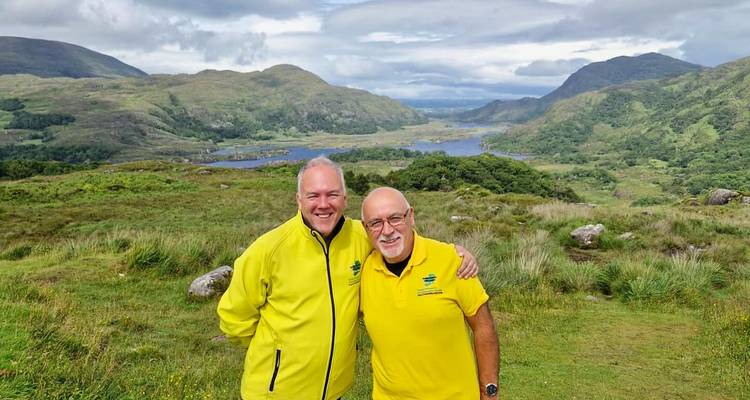 Deux hommes en vestes jaune vif sourient devant une vallée irlandaise luxuriante et des lacs