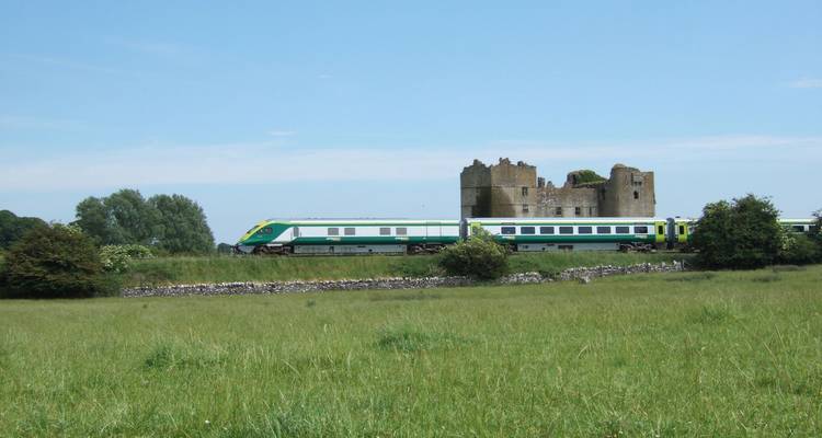 Un train d'Irish Rail passe devant un château en ruines à travers un champ vert sous un ciel dégagé