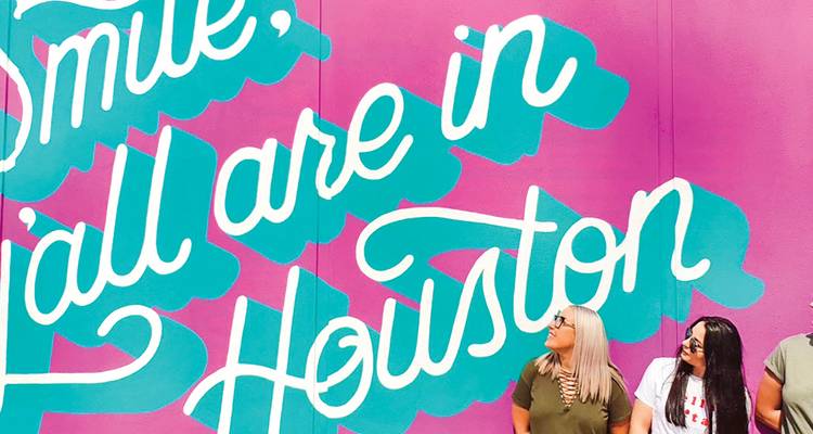 Friends look up at a colourful wall mural that reads "Smile, y'all are in Houston".