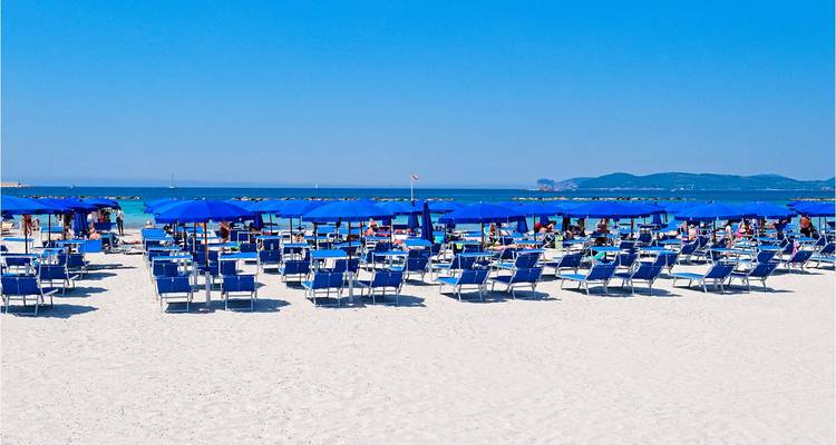 Playa de arena blanca y soleada con filas de tumbonas azules y sombrillas frente a un mar turquesa con colinas a lo lejos.