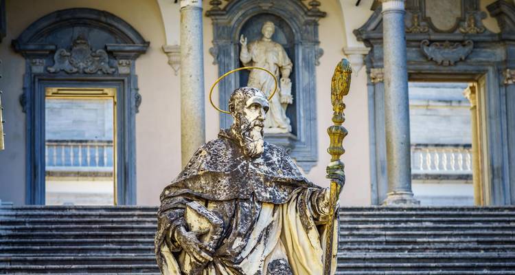 Estatua de mármol desgastada de un santo con halo se encuentra frente a los escalones de un gran monasterio.