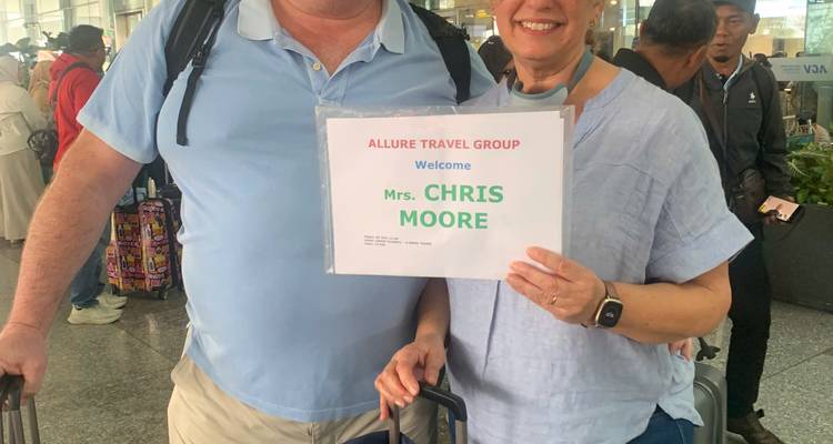 Twee reizigers in een aankomsthal van een luchthaven die een gedrukt welkomstbord en bagage vasthouden.