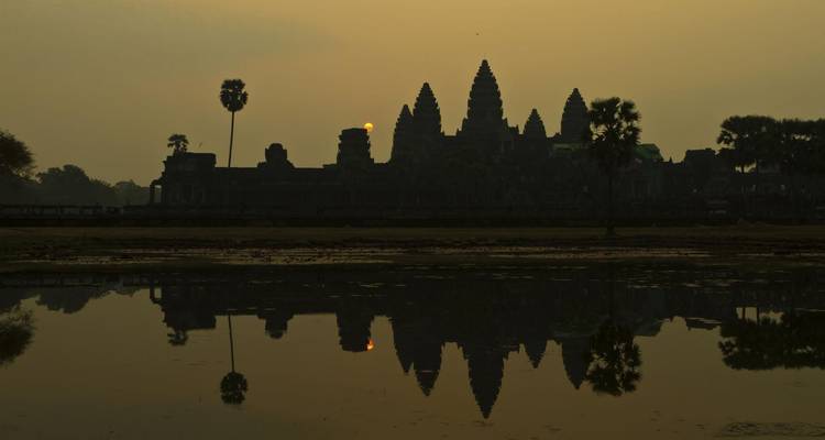 Silueta de Angkor Wat y palmeras reflejadas en agua tranquila al amanecer.