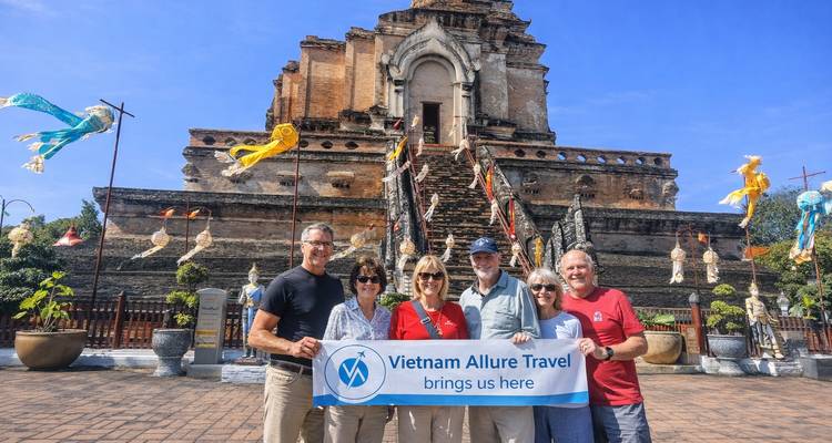 Grupo turístico sonriente sosteniendo una pancarta de Vietnam Allure Travel frente a las ruinas del templo Wat Chedi Luang.