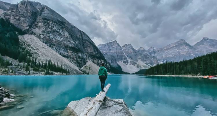 Person steht auf einem Baumstamm an einem blauen Alpensee mit Bergen.