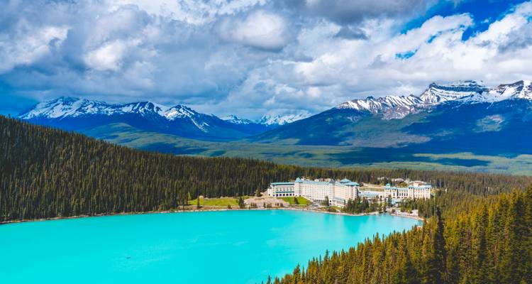 Hotel an einem türkisfarbenen See mit schneebedeckten Bergen in der Ferne.