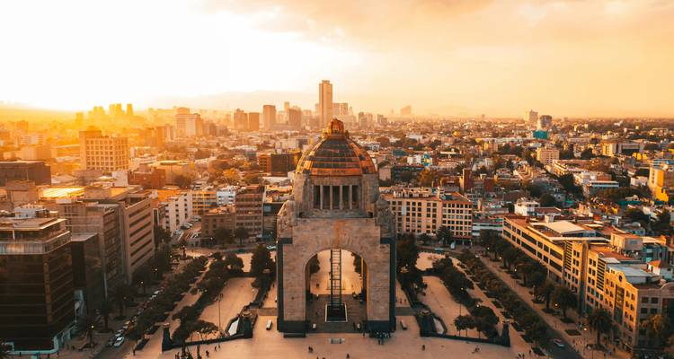 Vista aérea del Monumento a la Revolución rodeado por el skyline de la Ciudad de México bajo la cálida luz del atardecer.