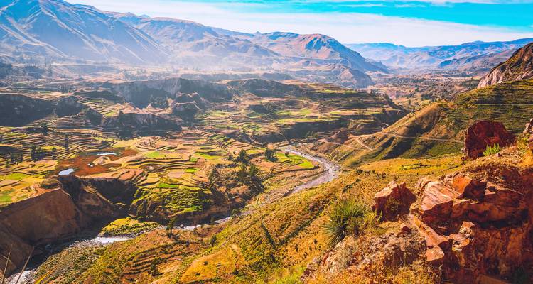 Uitgestrekt terrashellingslandschap met een kronkelende rivier en kleurrijke landbouwgronden omgeven door de Andes-bergen.