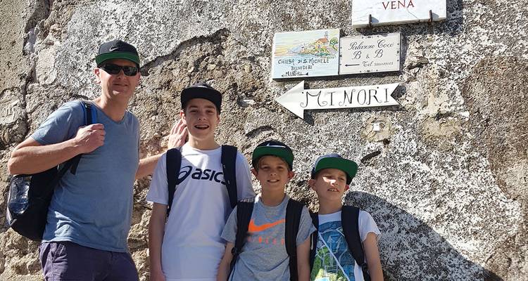 Un padre y tres niños sonríen junto a carteles pintados a mano que señalan hacia el pueblo de Minori en una pared rústica.