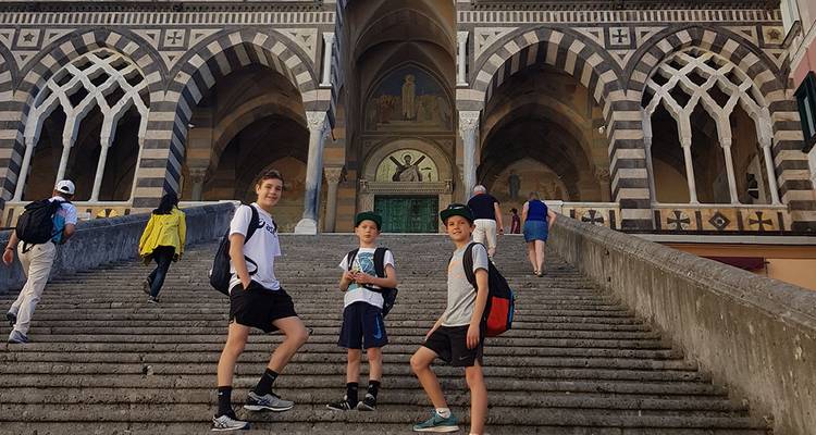Los adolescentes posan en la amplia escalinata de piedra de una catedral de fachada rayada en un pueblo costero italiano.