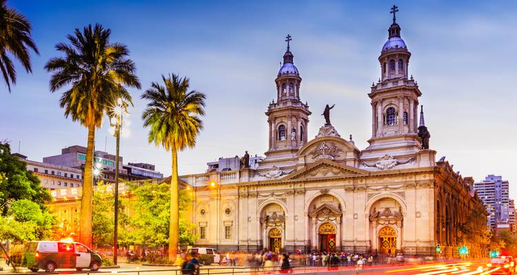 Catedral Metropolitana de Santiago iluminada al anochecer con palmeras y desenfoque de movimiento del tráfico que pasa.