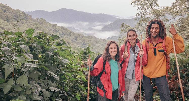 Tres excursionistas sonrientes con chaquetas rojas posan en un sendero selvático frondoso con colinas brumosas al fondo