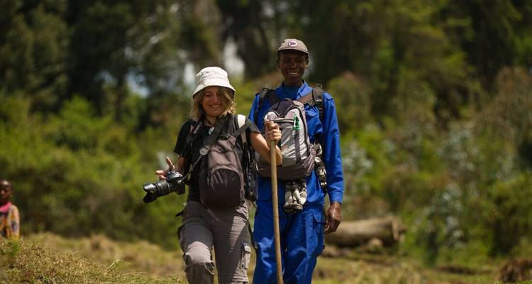 Viajera con cámara camina junto a guía local por un sendero soleado y cubierto de hierba