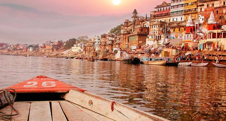 Uitzicht vanuit een houten boot die de kleurrijke ghats van Varanasi nadert, gebaad in zacht ochtendlicht over de Ganges.