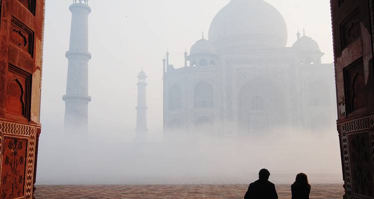 Silhouetten van twee bezoekers omlijst door Mogolbogen terwijl mist het Taj Mahal bij dageraad omringt.