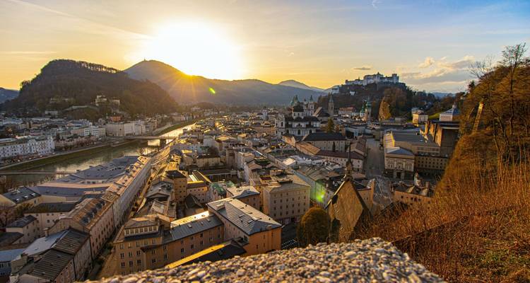 Cityscape at sunset with river and castle on a hill.