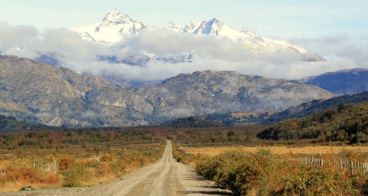 Un largo camino de grava conduce hacia las cumbres andinas cubiertas de nieve bajo nubes dispersas.