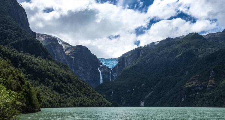 Lago turquesa rodeado de empinadas laderas boscosas con un glaciar a lo lejos que desciende entre los picos.