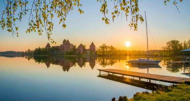 Trakai Island Castle en een zeilboot weerspiegeld in een spiegelglad meer bij zonsopgang met overhangende berkenbladeren.