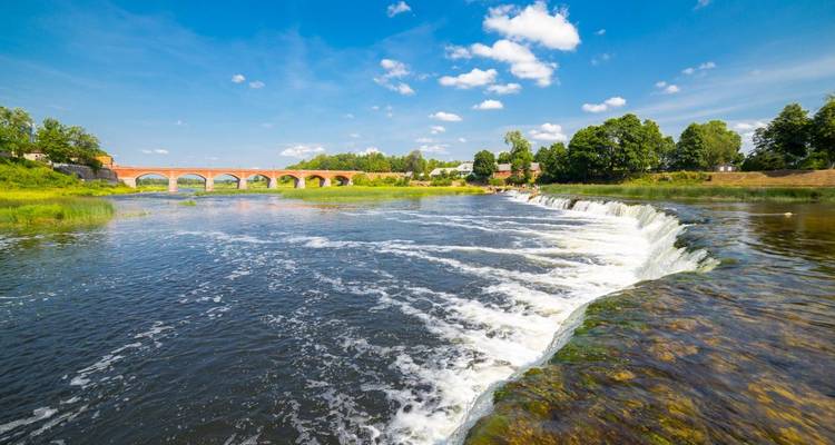 Brede rivier waterval en historische rode bakstenen brug omlijst door groene bomen onder een helderblauwe hemel.