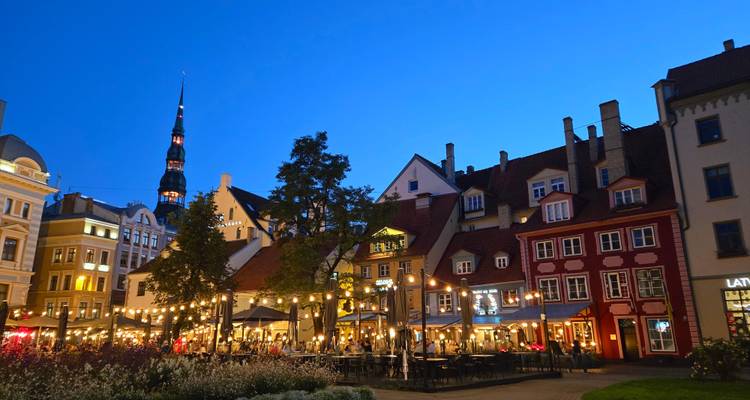 Avondaanzicht van Riga's Oude Stad met verlichte cafés, slingerlampen en historische torenspitsen tegen een diepblauwe hemel.