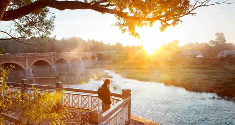 Reiziger geniet van zonsopgang bij een mistige rivier en historische bakstenen brug, gouden stralen verlichten het tafereel.